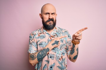 Handsome bald man with beard and tattoo wearing casual floral shirt over pink background Pointing...
