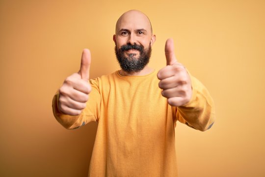 Handsome bald man with beard wearing casual sweater standing over yellow background approving doing positive gesture with hand, thumbs up smiling and happy for success. Winner gesture.