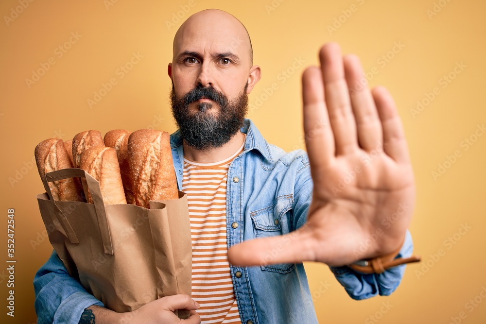 Sticker Handsome bald man with beard holding paper bag with bread over yellow background with open hand doing stop sign with serious and confident expression, defense gesture