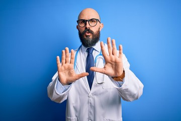 Handsome bald doctor man with beard wearing glasses and stethoscope over blue background Moving away hands palms showing refusal and denial with afraid and disgusting expression. Stop and forbidden.