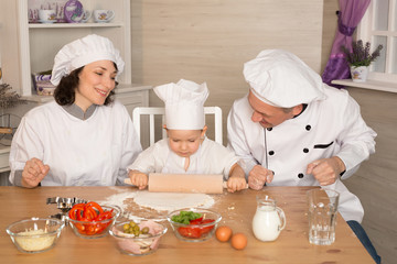 Mom, dad and little son cook together in quarantine. The family laughs and cooks pizza in cookery suits. A small child rolls out the dough with a rolling pin.