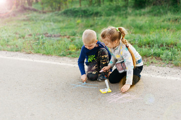Fototapeta premium Beautiful boy and girl blond brother and sister draw on the pavement with colored chalk. International Children's Day.