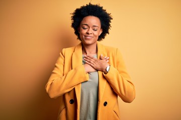 Young beautiful African American afro businesswoman with curly hair wearing yellow jacket smiling with hands on chest with closed eyes and grateful gesture on face. Health concept.