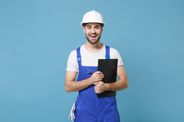 Funny young man in coveralls protective helmet hardhat hold clipboard with papers document isolated on blue wall background. Instruments accessories for renovation apartment room. Repair home concept.