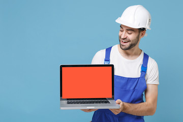 Funny man in coveralls protective helmet hardhat hold laptop computer with blank empty screen isolated on blue background. Instruments accessories for renovation apartment room. Repair home concept.