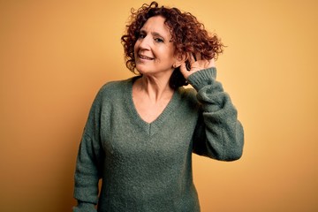 Middle age beautiful curly hair woman wearing casual sweater over isolated yellow background smiling with hand over ear listening an hearing to rumor or gossip. Deafness concept.