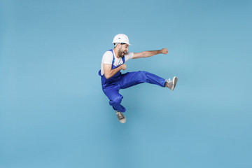 Fototapeta na wymiar Funny young man in coveralls protective helmet hardhat isolated on blue background in studio. Instruments accessories for renovation apartment room. Repair home concept. Having fun, jumping fighting.