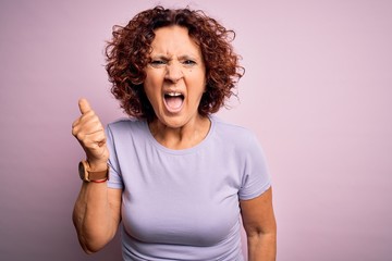 Middle age beautiful curly hair woman wearing casual t-shirt over isolated pink background angry and mad raising fist frustrated and furious while shouting with anger. Rage and aggressive concept.