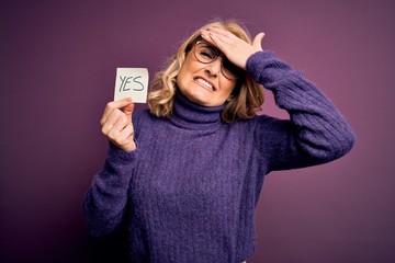 Middle age beautiful blonde woman wearing glasses holding reminder paper with yes message stressed...
