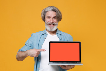 Funny elderly gray-haired mustache bearded man in casual blue shirt posing isolated on yellow background. People lifestyle concept. Pointing index finger on laptop pc computer with blank empty screen.