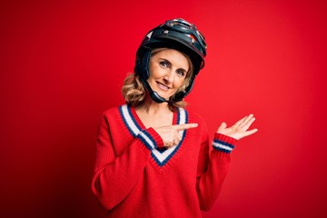 Middle age beautiful blonde motorcyclist woman wearing moto helmet over red background amazed and smiling to the camera while presenting with hand and pointing with finger.