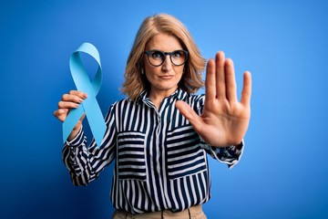 Middle age beautiful blonde woman wearing glasses holding blue cancer ribbon with open hand doing...