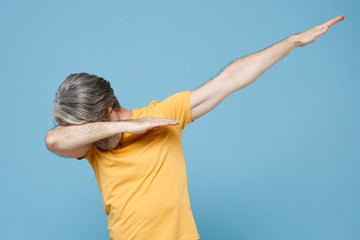 Elderly gray-haired mustache bearded man in casual yellow t-shirt posing isolated on pastel blue background studio portrait. People lifestyle concept. Mock up copy space. Showing DAB dance gesture.