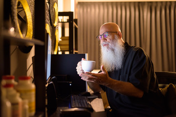 Happy mature bald bearded man drinking coffee while video calling at work from home late at night