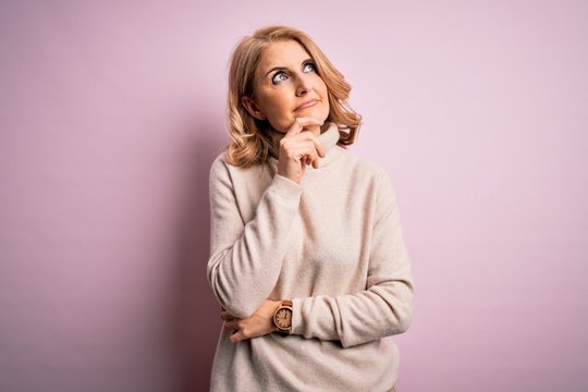 Middle age beautiful blonde woman wearing casual turtleneck sweater over pink background with hand on chin thinking about question, pensive expression. Smiling with thoughtful face. Doubt concept.
