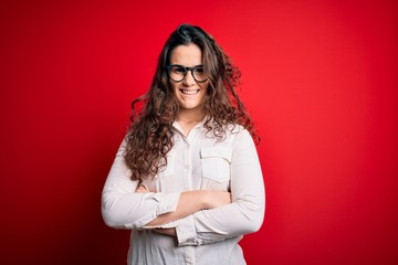 Young beautiful woman with curly hair wearing shirt and glasses over red background happy face smiling with crossed arms looking at the camera. Positive person.