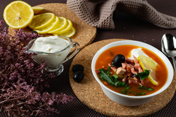 Soup Solyanka is a combined meat soup with smoked meat , various types of meat and tomato paste, serving dishes with sour cream, lemon and fresh herbs. Black olives on the table and a sprig of fresh f