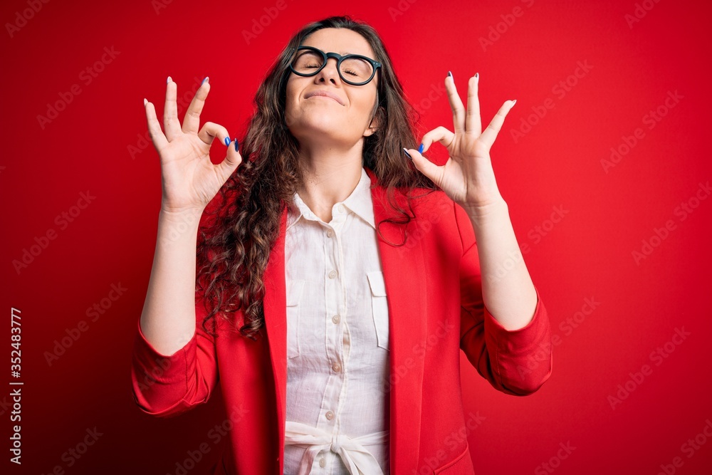 Wall mural young beautiful woman with curly hair wearing jacket and glasses over red background relax and smili