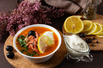 Soup Solyanka is a combined meat soup with smoked meat , various types of meat and tomato paste, serving dishes with sour cream, lemon and fresh herbs. Black olives on the table and a sprig of fresh f