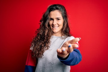 Young beautiful woman with curly hair wearing casual sweatshirt over isolated red background smiling friendly offering handshake as greeting and welcoming. Successful business.