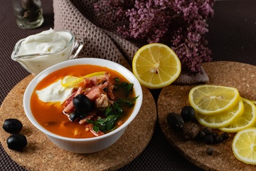 Soup Solyanka is a combined meat soup with smoked meat , various types of meat and tomato paste, serving dishes with sour cream, lemon and fresh herbs. Black olives on the table and a sprig of fresh f