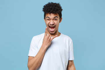 Fototapeta na wymiar Crazy young african american guy in casual white t-shirt posing isolated on pastel blue background. People lifestyle concept. Mock up copy space. Depicting heavy metal rock sign, horns up gesture.