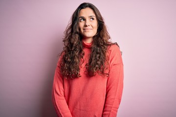 Young beautiful woman with curly hair wearing turtleneck sweater over pink background smiling looking to the side and staring away thinking.