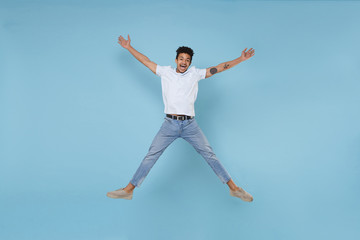 Fototapeta na wymiar Excited young african american guy in casual white t-shirt posing isolated on pastel blue background studio portrait. People lifestyle concept. Mock up copy space. Jumping, spreading hands and legs.