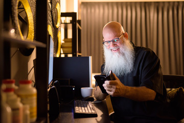 Happy mature bald bearded man using phone while working overtime at home late at night