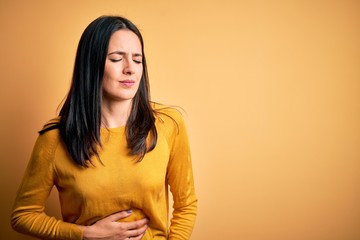 Obraz premium Young brunette woman with blue eyes wearing casual sweater over yellow background with hand on stomach because indigestion, painful illness feeling unwell. Ache concept.