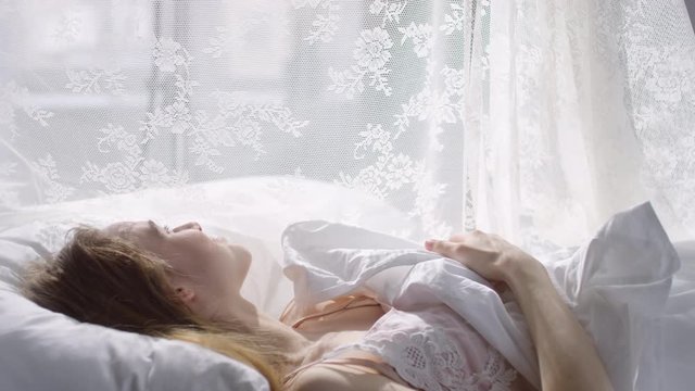  Slowmo of happy young woman in nightdress lying in bed on serene morning and smiling while enjoying sunlight shining through lace canopy