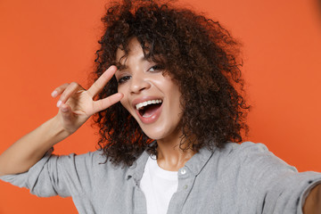Close up of cheerful african american woman in gray casual clothes isolated on orange background. People lifestyle concept. Mock up copy space. Doing selfie shot on mobile phone, showing victory sign.