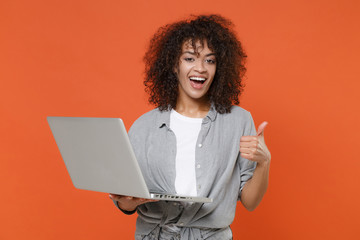 Excited young african american woman girl in gray casual clothes isolated on orange wall background in studio. People lifestyle concept. Mock up copy space. Hold laptop pc computer, showing thumb up.