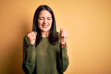 Young brunette woman with blue eyes wearing green casual sweater over yellow background excited for success with arms raised and eyes closed celebrating victory smiling. Winner concept.