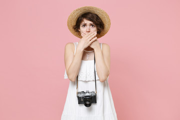 Upset tourist young girl in summer white dress hat with photo camera isolated on pink background in studio. Traveling to travel weekends getaway. Air flight journey concept. Covering mouth with hands.