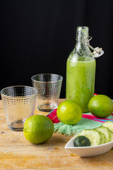 Top view of bottle with cucumber juice, limes, empty glasses and cucumber slices in bowl, on wooden table and black background, horizontal