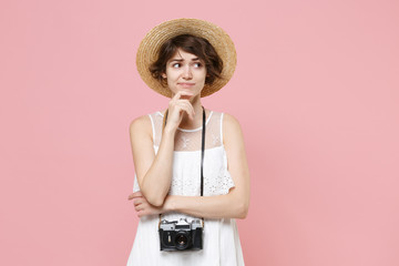 Pensive confused young tourist girl in summer white dress hat with photo camera isolated on pink...