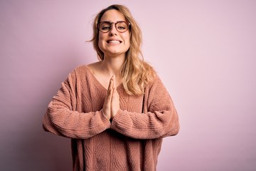 Young beautiful blonde woman wearing casual sweater and glasses over pink background praying with hands together asking for forgiveness smiling confident.