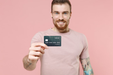 Smiling young bearded tattooed man guy in pastel casual t-shirt posing isolated on pink wall background studio portrait. People emotions lifestyle concept. Mock up copy space. Hold credit bank card.