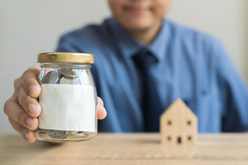 Money savings concepts  wood house models with blur Men are showing Glass bottel for saving money with blank label and blur wooden house on wooden table with blur background.