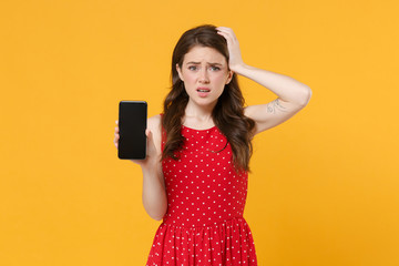 Naklejka premium Worried young brunette woman girl in red summer dress posing isolated on yellow background. People lifestyle concept. Mock up copy space. Hold mobile phone with blank empty screen put hand on head.