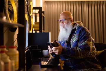 Mature bald bearded hipster man using phone while working overtime at home late at night