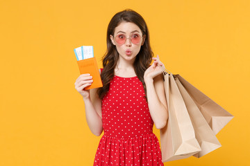 Amazed young brunette woman girl in red summer dress eyeglasses posing isolated on yellow background studio. People lifestyle concept. Hold package bag with purchases after shopping passport tickets.