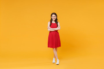 Smiling young brunette woman girl in red summer dress posing isolated on yellow wall background studio portrait. People sincere emotions lifestyle concept. Mock up copy space. Holding hands crossed.