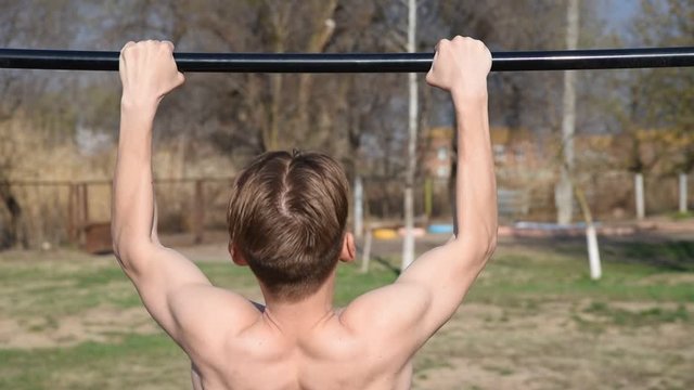 a young man with a naked torso pulls up on a horizontal bar on a sports field.Sport, street workout concept