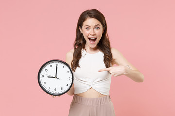 Excited young brunette woman girl in light casual clothes posing isolated on pastel pink wall background studio portrait. People lifestyle concept. Mock up copy space. Pointing index finger on clock.