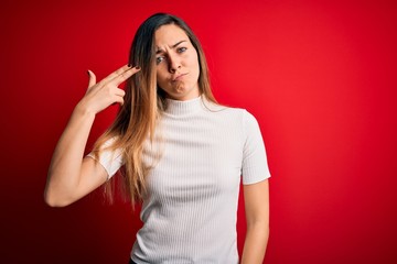 Beautiful blonde woman with blue eyes wearing casual white t-shirt over red background Shooting and killing oneself pointing hand and fingers to head like gun, suicide gesture.
