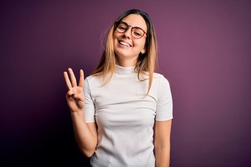 Young beautiful blonde woman with blue eyes wearing casual t-shirt over purple background showing and pointing up with fingers number three while smiling confident and happy.