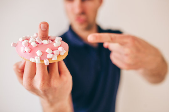 Young man isolated over background. Blurred and defocused picture. Guy show fuck sign and hold pink tasty delicious donut on it. Point on dessert with another finger.