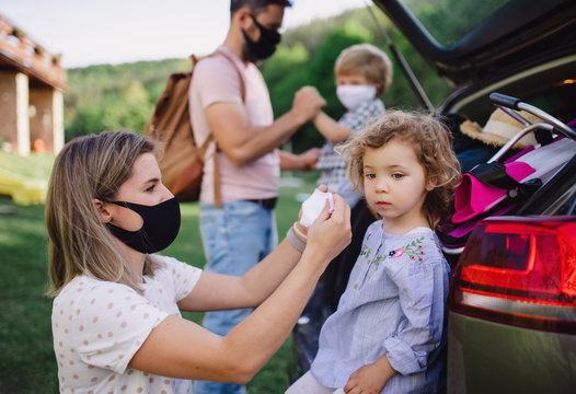 Family With Two Small Children Loading Car For Trip In Countryside, Wearing Face Masks.